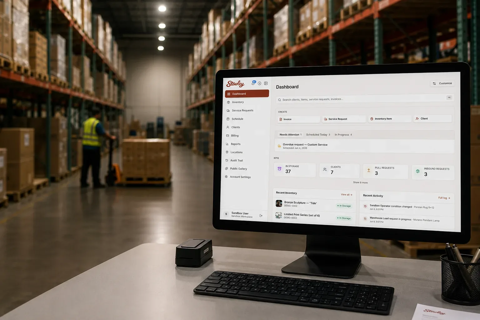 Multi-client warehouse software dashboard on a monitor inside a boutique 3PL facility, with a worker moving pallets between aisles of client inventory